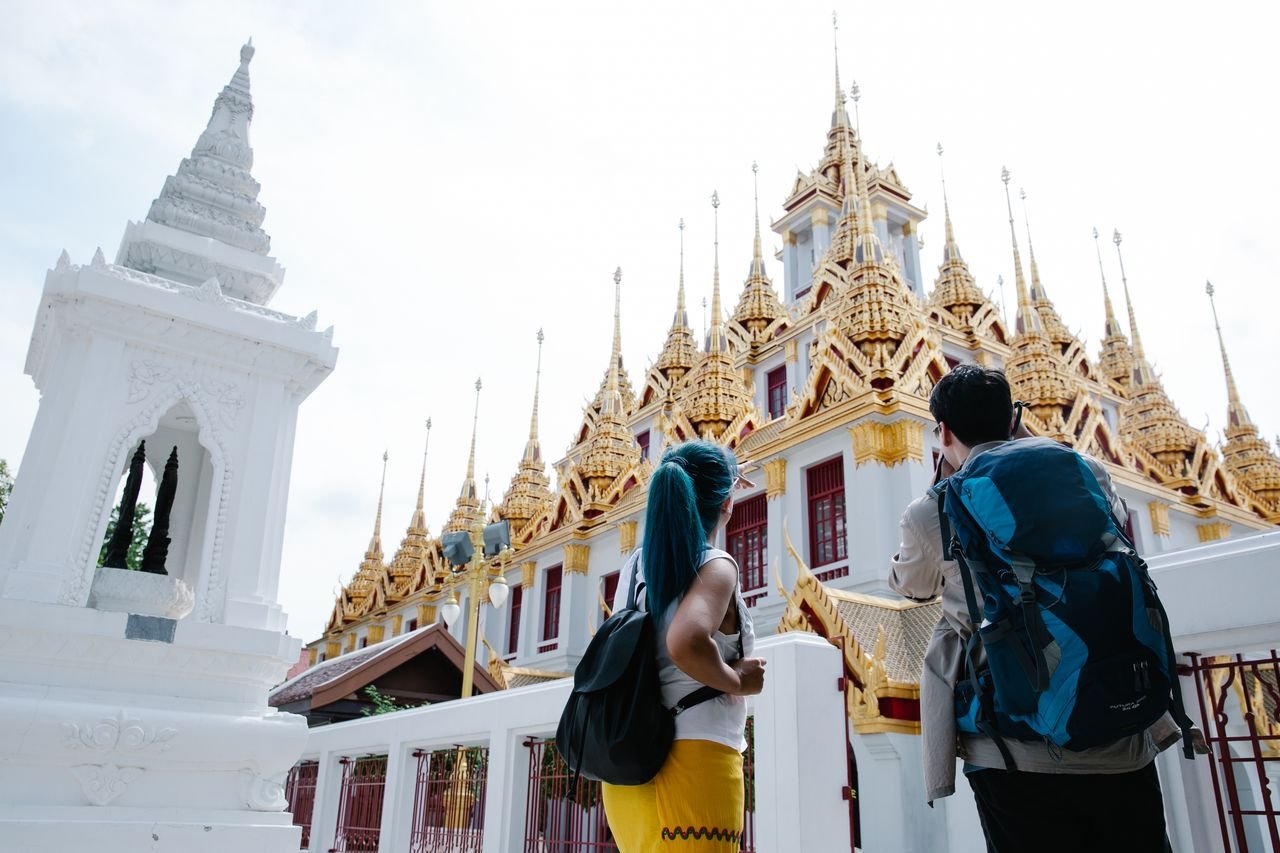 Two tourists with backpacks admire the ornate facade of a traditional Thai temple.